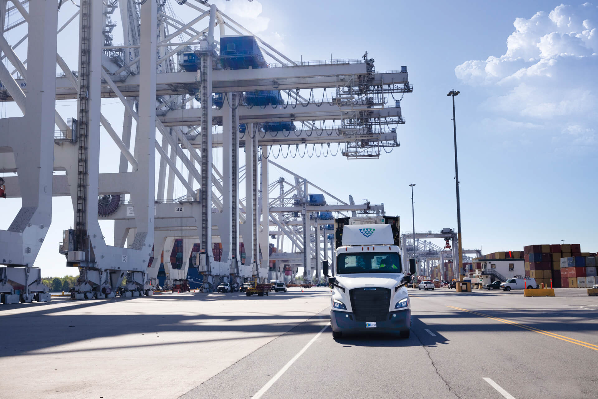 A Lineage Logistics truck drives through a busy port terminal, surrounded by large shipping cranes, cargo containers, and port infrastructure under a clear blue sky.