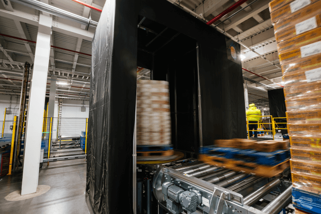 Lineage Eye and a high-speed conveyor system in a cold storage warehouse scanning pallets using Lineage Eye automation. A worker in a high-visibility vest monitors operations.