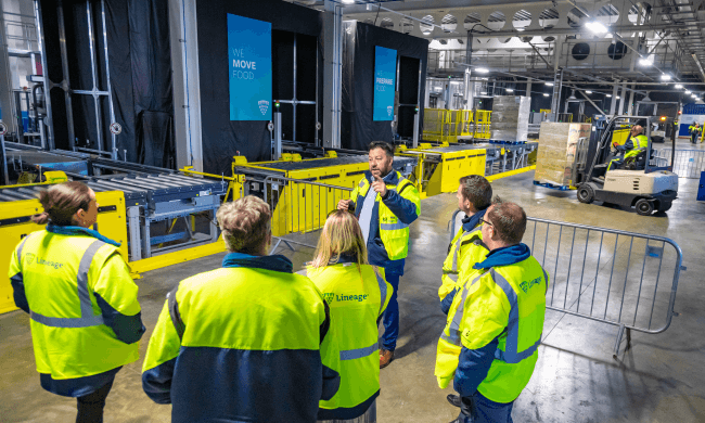 A group of Lineage team members in high-visibility vests discussing warehouse automation near the Lineage Eye system, with conveyor belts and forklifts in the background.