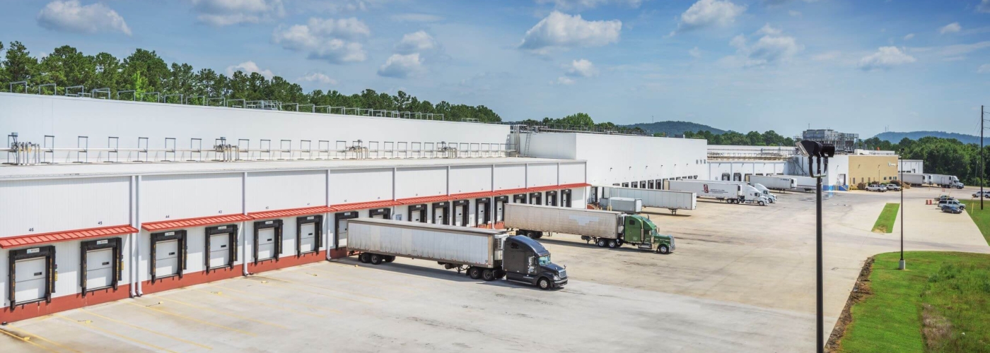 A view of Lineage's cold storage warehouse in Attalla, Alabama, showcasing multiple loading docks with semi-trucks parked for deliveries. The large white facility is surrounded by an expansive concrete lot and green landscaping under a bright blue sky with scattered clouds.