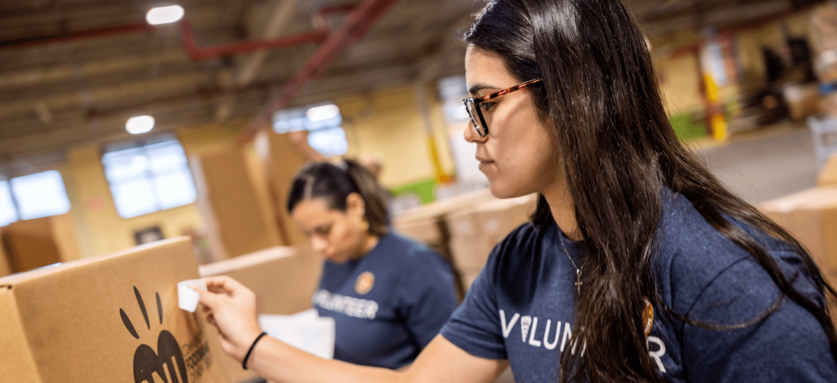 Two Lineage volunteers, wearing blue "Volunteer" shirts, pack and label boxes of food inside a warehouse, preparing food for donation to food insecure communities.