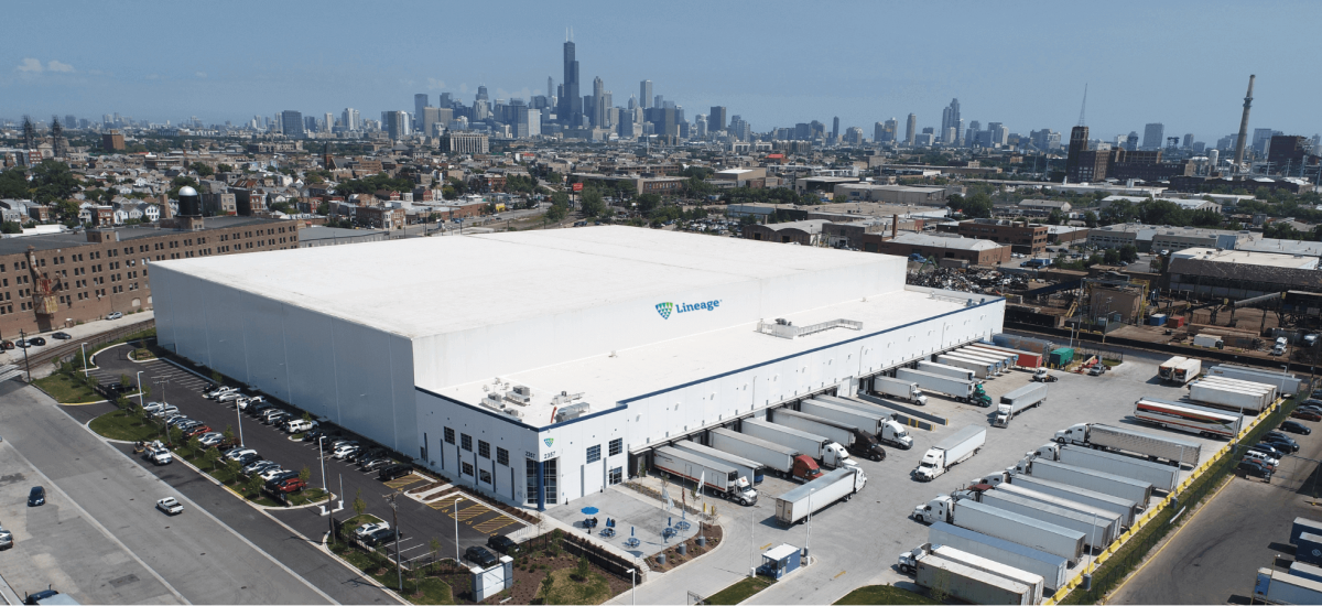 Aerial view of Lineage’s cold storage warehouse in Chicago, with the city skyline in the background. Numerous trucks are docked at loading bays, showcasing the facility’s capacity for large-scale distribution. The warehouse is a white building with the Lineage logo, surrounded by parking areas and greenery.