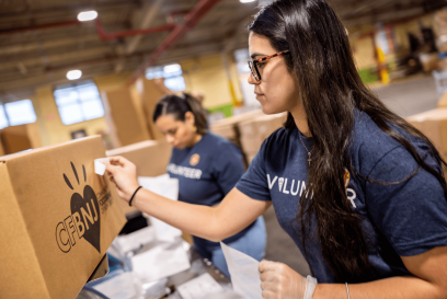 Two Lineage volunteers, wearing blue "Volunteer" shirts, pack and label boxes of food inside a warehouse, preparing food for donation to food insecure communities.